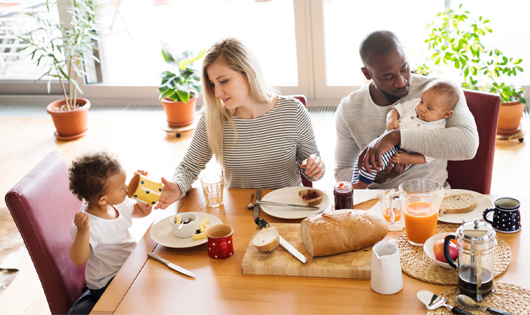 Eine Familie mit zwei kleinen Kindern sitzt um einen Esstisch und frühstückt.