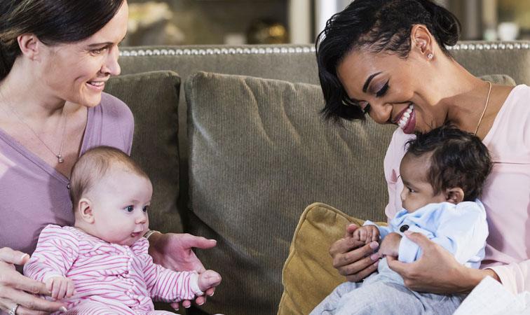 Zwei Mütter sitzen einander zugewandt auf einem Sofa und halten Ihre Babys auf dem Schoß